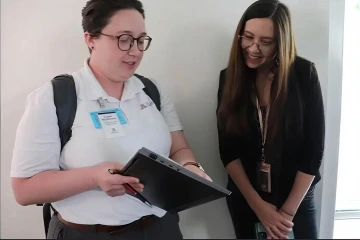 Two people stand together. One person is holding a piece of paper and a laptop and the other person is looking at it.