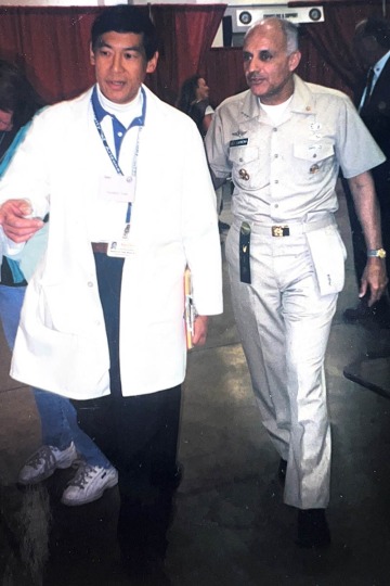 A young Asian man wearing a white coat walks beside a man wearing a military uniform.