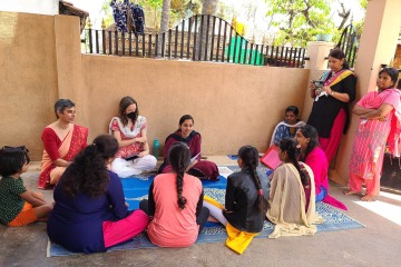 Group of women wearing Indian saris sit in a circle having a discussion. There are two women standing off to the side, also wearing saris.