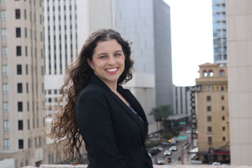 Woman with long, curly, brown hair stands on top of a building with taller buildings in the background.