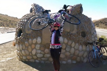 Woman wearing a black, white and pink bicycling outfit and helmet holds up a bicycle over her head. She is standing in front of a large, decorative rock.