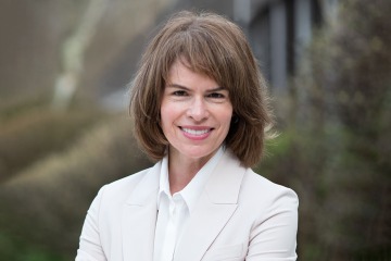 Portrait of a woman with shoulder length brown hair and bangs smiling. She is waring a light colored blazer and white dress shirt.