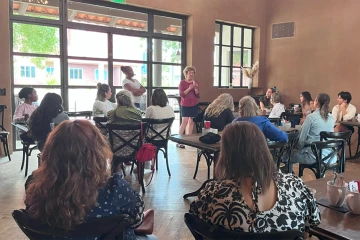 Group of people sit together at various tables in a cafe as a standing women speaks into a microphone.