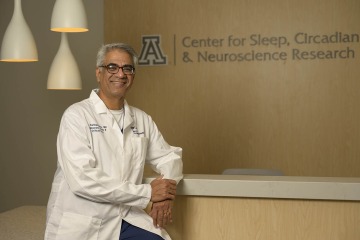 An brown-skinned man with gray hair and glasses and wearing a white coat leans against a counter. The words Center for Sleep, Circadian and Neuroscience Research can be seen on signage behind in.