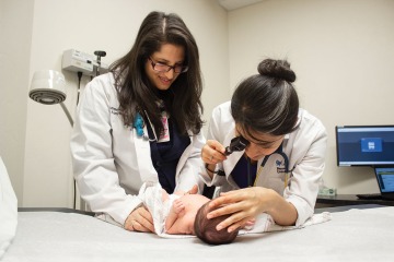 Two female doctors wearing white coats evaluate a newborn baby on a patient bed.