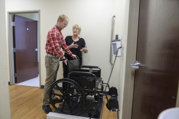 Female doctor wearing all black scrubs, shows a male medical student how to use a wheelchair scale in a clinic setting.