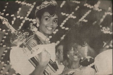 Newspaper photo of woman at a beauty pageant receiving a trophy and wearing a tiara.
