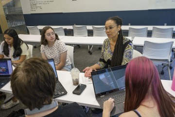 Female professor leads discussion in a classroom setting surrounded by college-age students.
