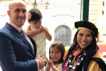 A family of four (a dad, two young daughters and a mom) stand together. The mom is wearing graduation regalia.