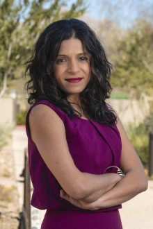 South Asian woman wearing a maroon dress stands outside with her arms crossed and a smiling face