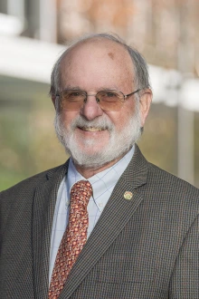 Older white man with white beard wearing a suit stands in front of a building