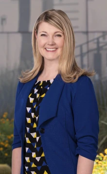 Woman with shoulder length blonde hair wearing a black, white, yellow and blue blouse with a blue blazer on top. She is smiling and standing outside.