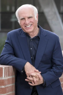 Man with grey hair wearing a blue suit leans against a half-high brick wall