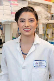 A young woman with dark hair wearing a white lab coat stands in a laboratory smiling.