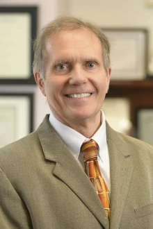 Portrait of Brian Erstad, an older white man with short hair smiling and wearing a tan suit and tie. 