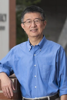 Portrait of Dr. Xinxin Ding standing outside leaning on a red brick wall. 