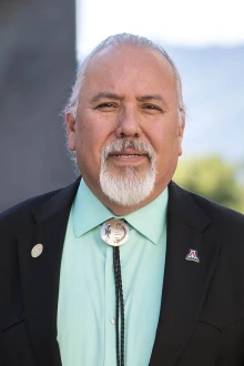 Portrait of Dr. Carlos Gonzales standing outsice in a dark suit coat and bolo tie.