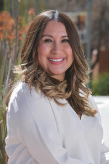 Portrait of a woman with long brown hair wearing a white shirt sits outside smiling.