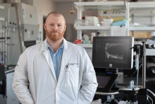 Light-skinned man with a red beard wearing a blue shirt and a white lab coat stands in a lab.