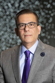 Portrait of a man wearing a grey suit, purple tie and glasses standing in front of lights in the Health Sciences Innovation Building Forum.