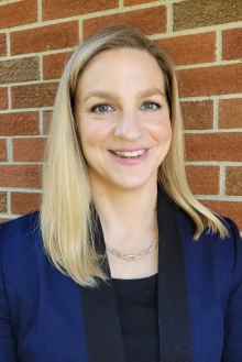 Portrait of professor Lindsay Bouchard wearing a blue jacket and smiling while standing in front of a brick wall. 