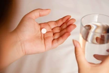 Open hand holding round white pill, other hand holding glass of water 