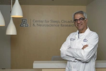 Portrait of Sai Parthasarathy, MD, in lab coat in front of U of A Health Sciences Sleep, Circadian & Neuroscience Research sign