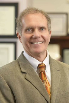 Portrait of Brian Erstad, an older white man with short hair smiling and wearing a tan suit and tie.