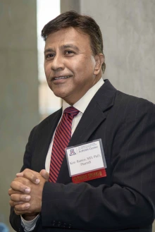 A man in a dark suit speaks into a microphone at a lectern; his hands are clasped in front of him.