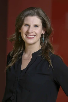 Portrait of professor Rina Fox smiling as she stands in front of a red wall with a large “A” on it.