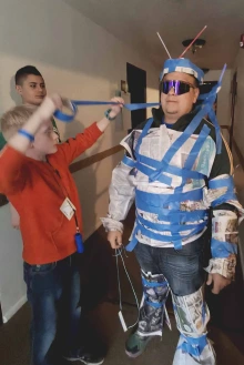 Two boys stand next to Alejandro Vazquez, and one is wrapping blue tape around Vazquez, who wears futuristic sunglasses and is covered in newspaper and tape as part of a costume. 