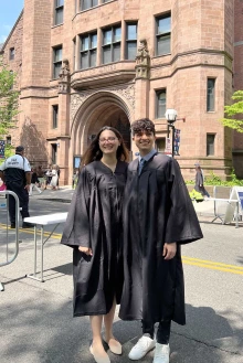 Jerry Ruvalcaba and Katie Simic, wearing black graduation gowns, stand side by side and smile in front of a large, brown stone Gothic building at Yale University.