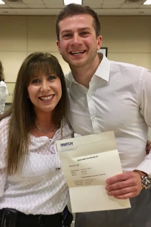 A woman and man stand together. The man is holding his Match Day letter. 