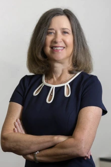 Photo of an older woman with grayish-brown hair, wearing a navy blue dress and standing in an office.