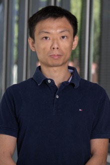 Asian man with dark brown hair and wearing a black t-shirt stands outside.