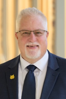 Portrait of an older white man with gray hair and beard wearing a navy blue suit and a gold University of Arizona Wildcat lapel pin,