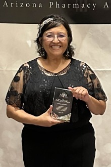 Woman with shoulder length dark hair and glasses wearing a black blouse and plants holds an award while smiling.