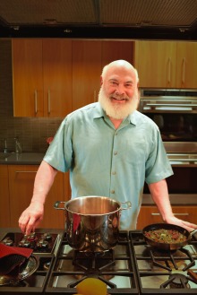 Man with grey beard stands in a kitchen in front of a large pot on the stove
