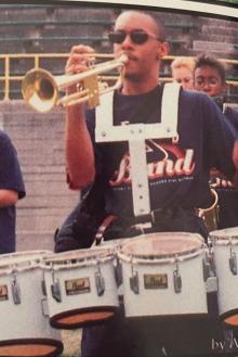 Picture of Michael D.L. Johnson in a high school yearbook photo, blowing a trumpet with drums strapped to him. 