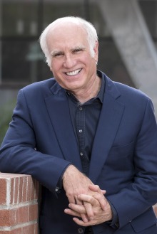Older man with white hair wearing a navy blue suit leans against a half-brick wall outside smiling.