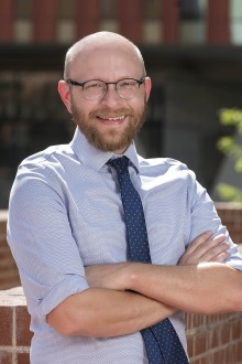 Portrait of Dr. Christopher Edwards outside wearing a grey shirt, blue tie with white polka dots and smiling.