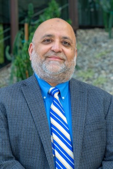 Dr. Shakaib Rehman smiling. He is wearing a blue dress shirt, grey suit jacket and blue and yellow tie.
