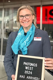 Dr. Jennifer Barton stands in front of a building holding a sign that reads, “Photonics West 2024 has arrived.”