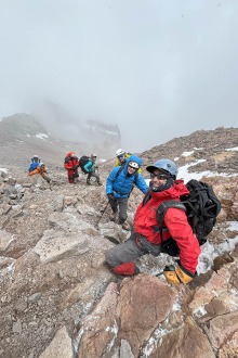Six people in parkas and climbing gear climb up Aconcagua with clouds in the background. 