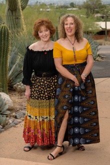 Two women stand next to each other in a desert setting. Both are wearing skirts and off the shoulder tops.