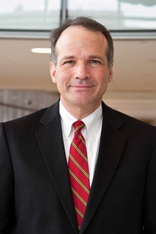 Portrait of man with grey and brown hair wearing a black suit, white dress shirt and red tie.