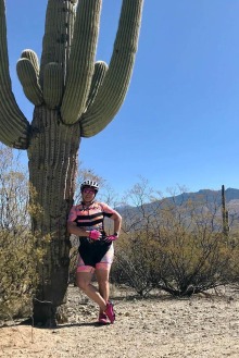 Woman wearing a pink and black bicycling outfit and helmet leaning against a saquaro cactus.