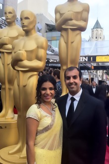 Dr. Rachna Shroff in a yellow saree and her husband Dr. Puneet Shroff stand in front of life-size Oscar trophies.